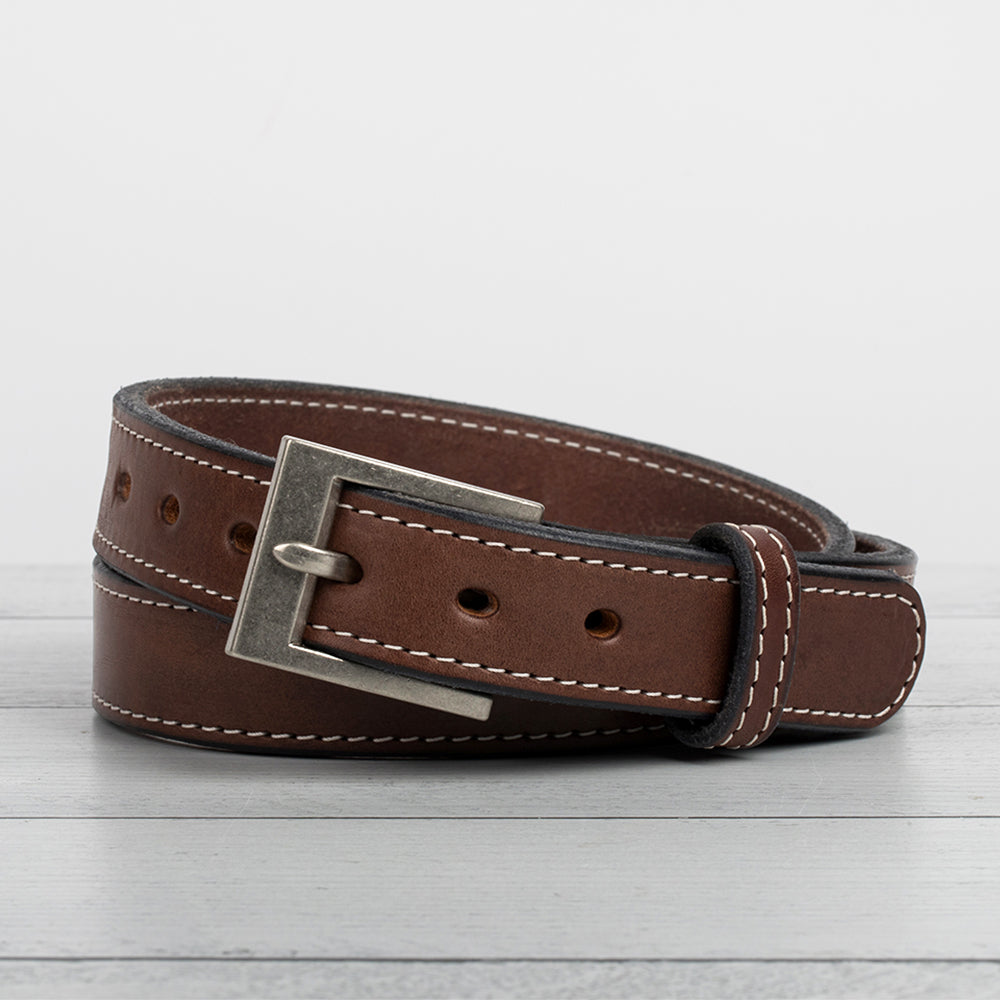 The Bonnie by Hanks Belts is a brown, full-grain vegetable tanned leather belt with white stitching and a silver rectangular buckle, coiled on light wood against a plain background.