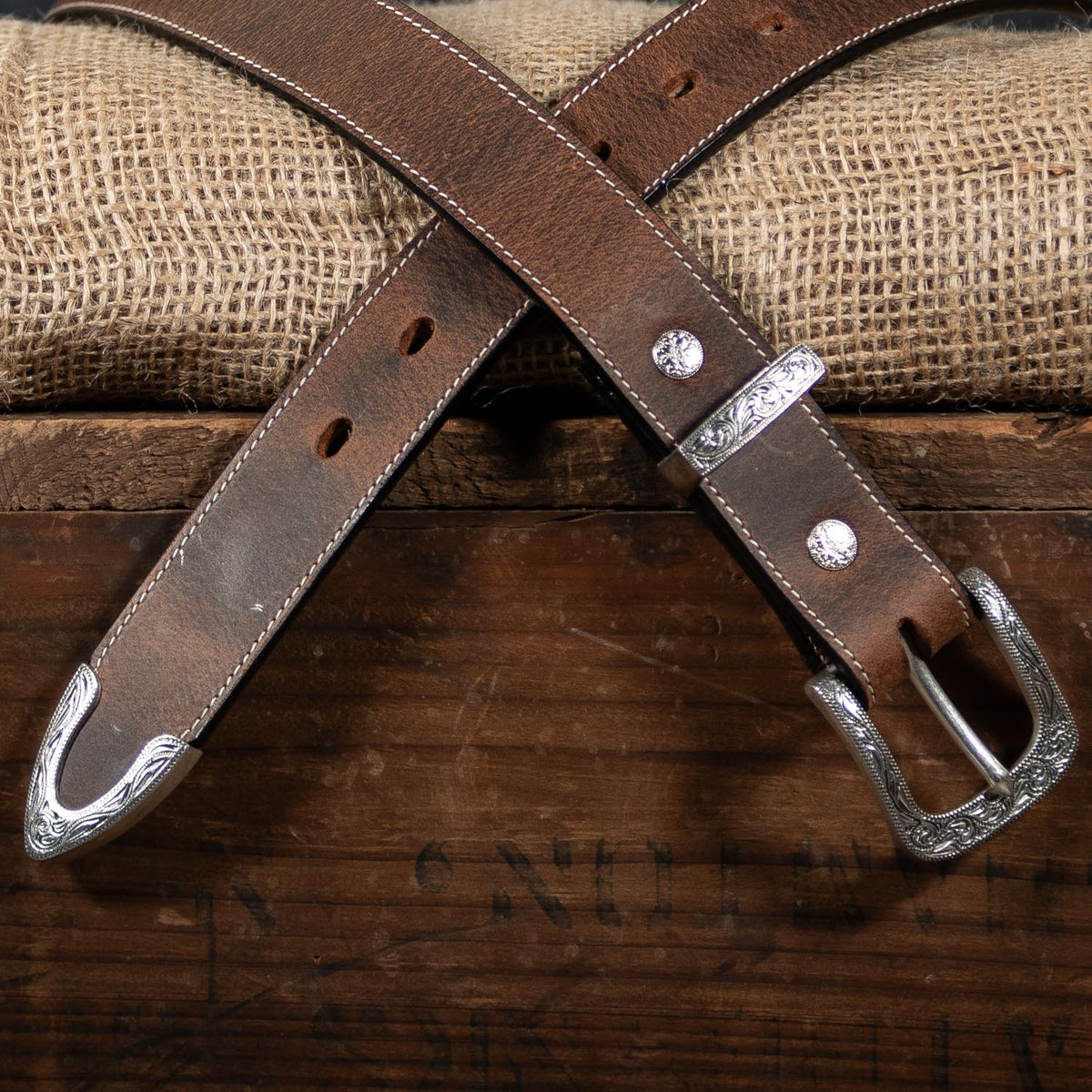 The Dakota Bison by Hanks Belts, crafted from North American bison leather with a silver floral buckle and tip, is displayed crosswise on a rustic wooden box, burlap sack in the background.
