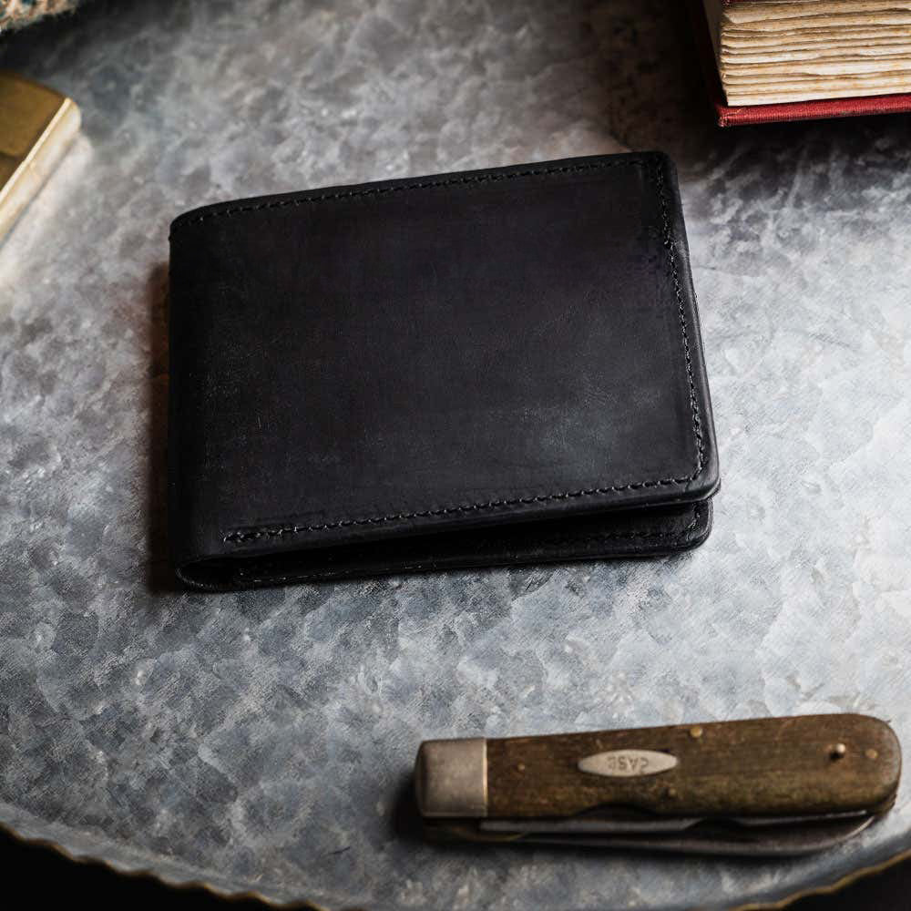 The Huron Premium Bifold with ID Window by Hanks Belts rests on a round, textured metal surface beside a closed pocketknife and several worn books.