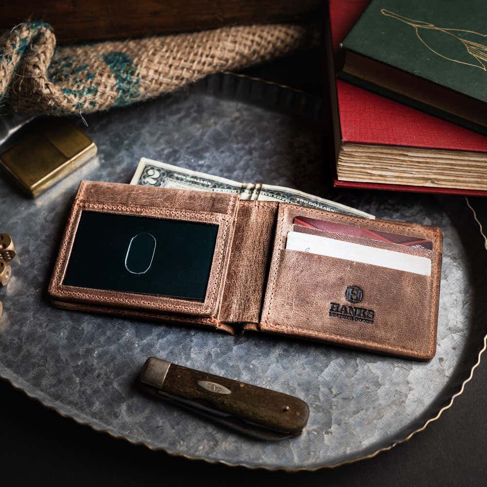 The Huron Premium Bifold with ID Window by Hanks Belts, crafted in the USA, sits open on a metal tray displaying cards, cash, and a black card. Nearby are a pocketknife, gold lighter, and red books for a vintage touch.