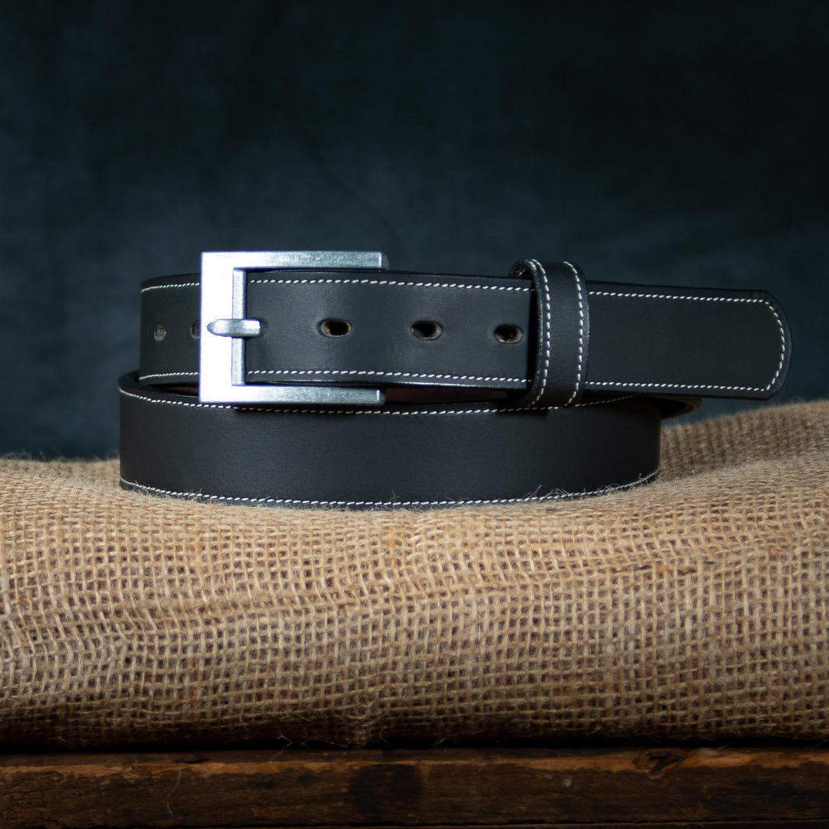 The Heritage Bison belt by Hanks Belts, crafted from black bison leather with white stitching and a silver buckle, is coiled neatly on burlap, highlighting its rugged style against a dark backdrop.