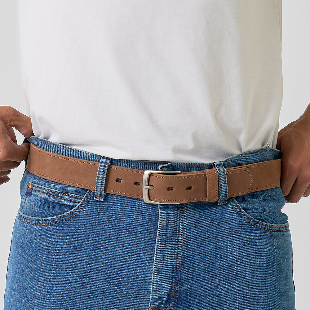 A person wearing a plain white T-shirt and blue jeans adjusts The Suede - 1.5" light brown Nubuck leather belt by Hanks Belts, featuring a silver buckle. Image is cropped to show only the waist and hands.