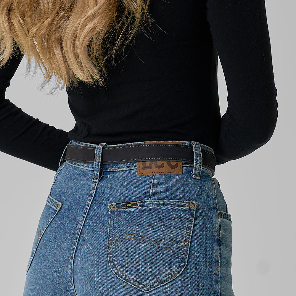 A person viewed from behind wears a black long-sleeve shirt, blue denim jeans, and The Lennon Suede belt by Hanks Belts. Their long, wavy blonde hair drapes over their shoulder.