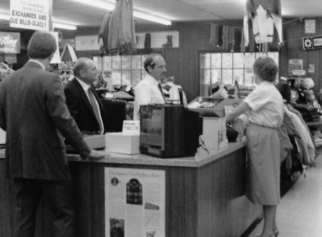 Early 70's. Vintage black and white photo of a store checkout area with customers and staff. Text below the image: As retail changed, so did Hank’s. Expanded offerings included casual clothing and footwear, all while still maintaining their focus on quality USA Made work wear and leather goods.