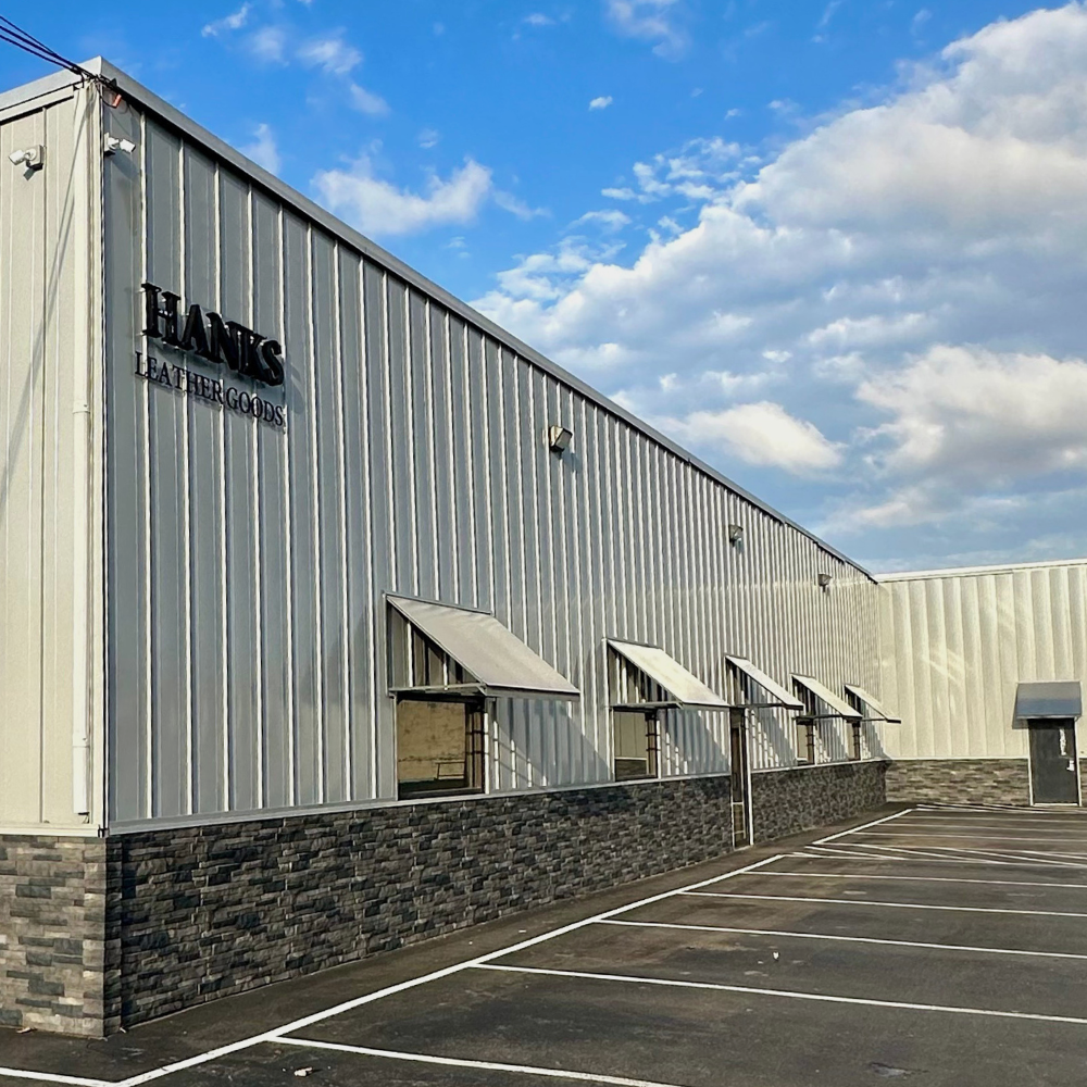 2011. Factory building with 'Hans Leather Goods' branding under a blue sky with clouds. Text below the image: Hank's moves into its new 30,000 square foot facility to accommodate the growing business. With this expansion, it provides the opportunity to expand our selection of leather products to now include wallets, dog accessories and more.