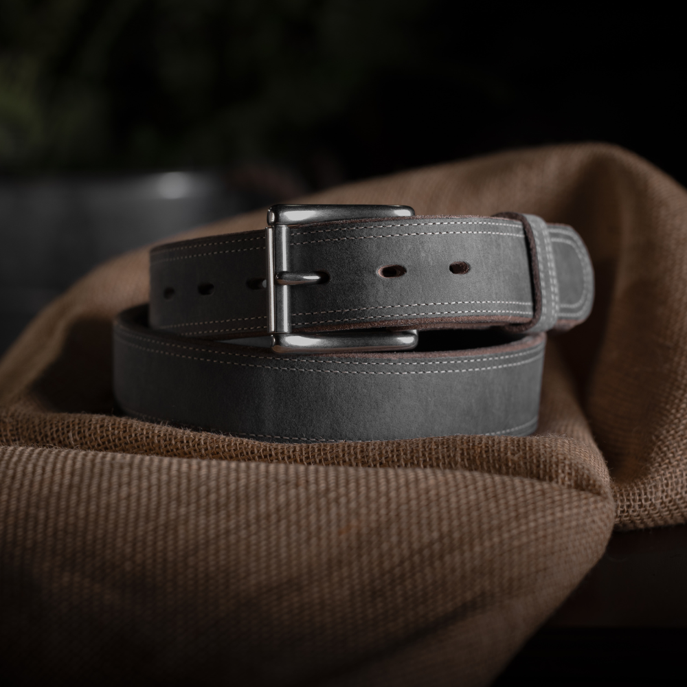 The Montana Bison Double Layer by Hanks Belts, featuring sturdy bison leather with white stitching and a stainless steel buckle, is coiled on brown textured fabric against a dark, blurred background.