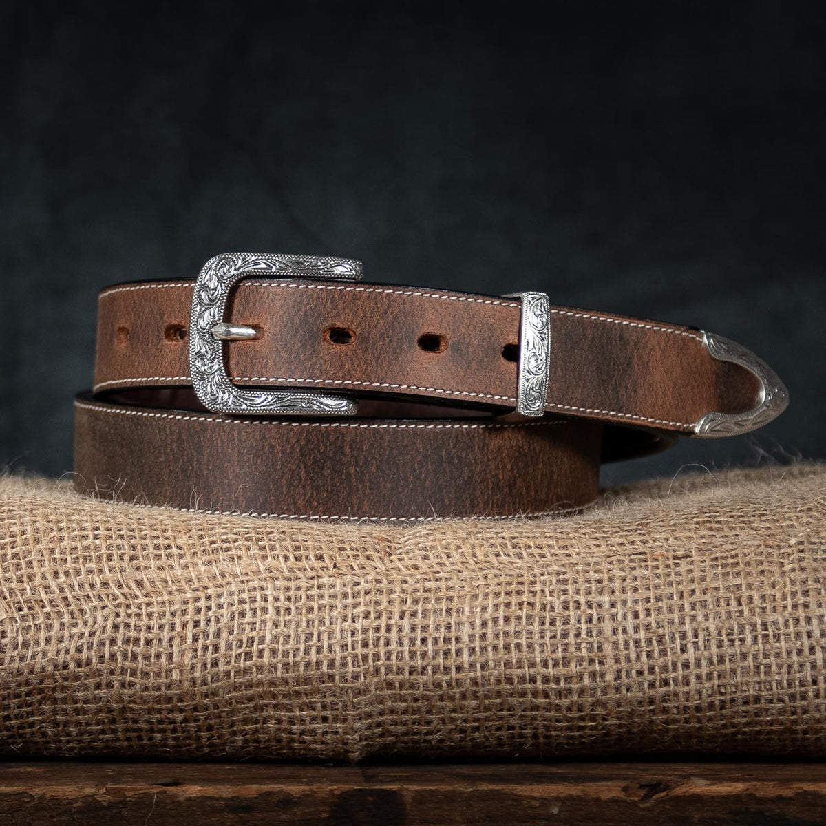 The Dakota Bison by Hanks Belts: a brown North American bison leather western belt with a silver floral buckle, tip, and loop, shown coiled on burlap against a dark background.