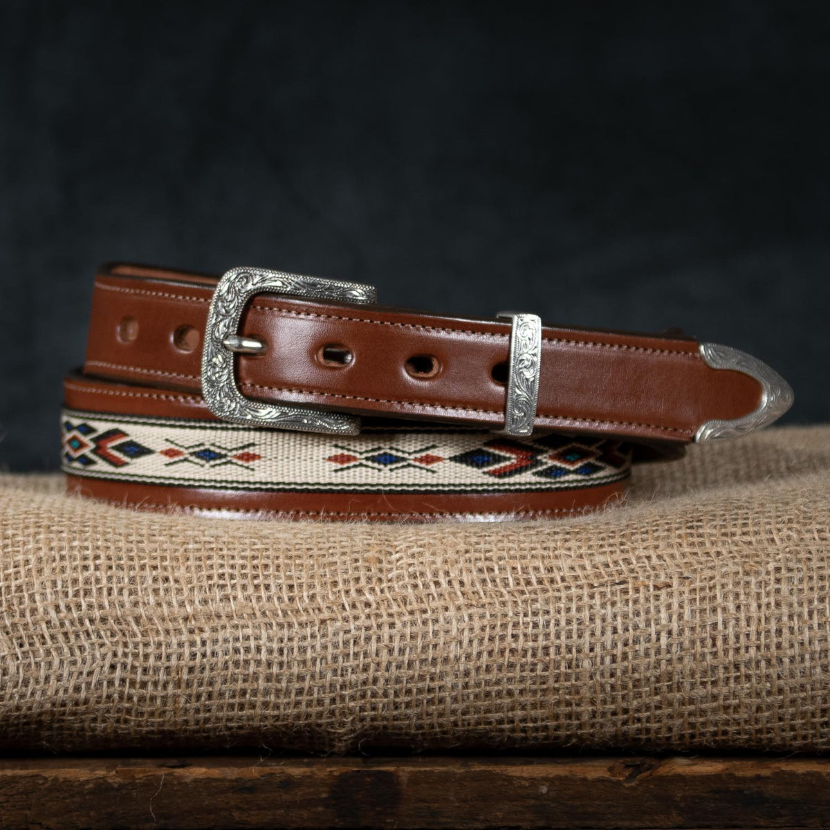 The Seneca Tapered by Hanks Belts is a brown English Bridle leather belt with a decorative red, navy, and cream woven pattern and an engraved silver buckle and tip, shown on burlap against a dark background.