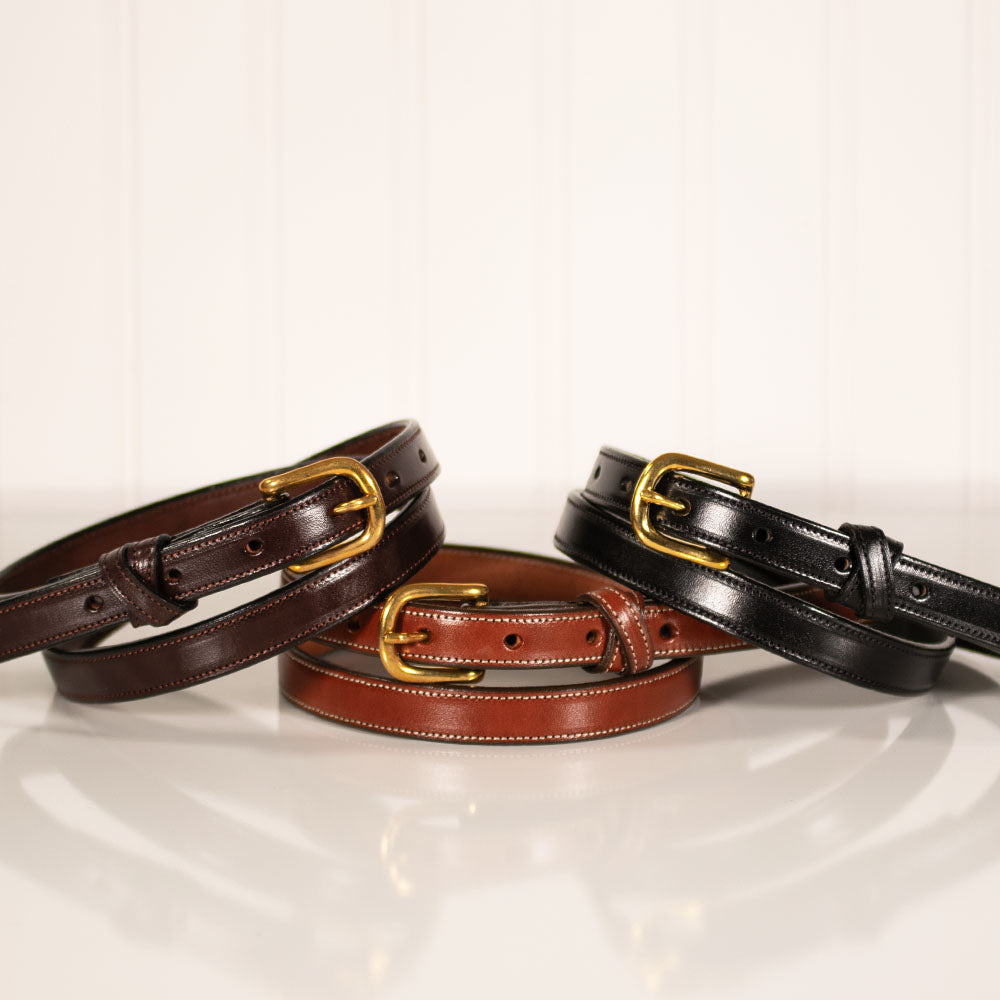 Three women's belts—The Dixie by Hanks Belts in dark brown, tan, and black English Bridle leather with gold buckles—are neatly coiled side by side on a reflective white surface against a white panel background.