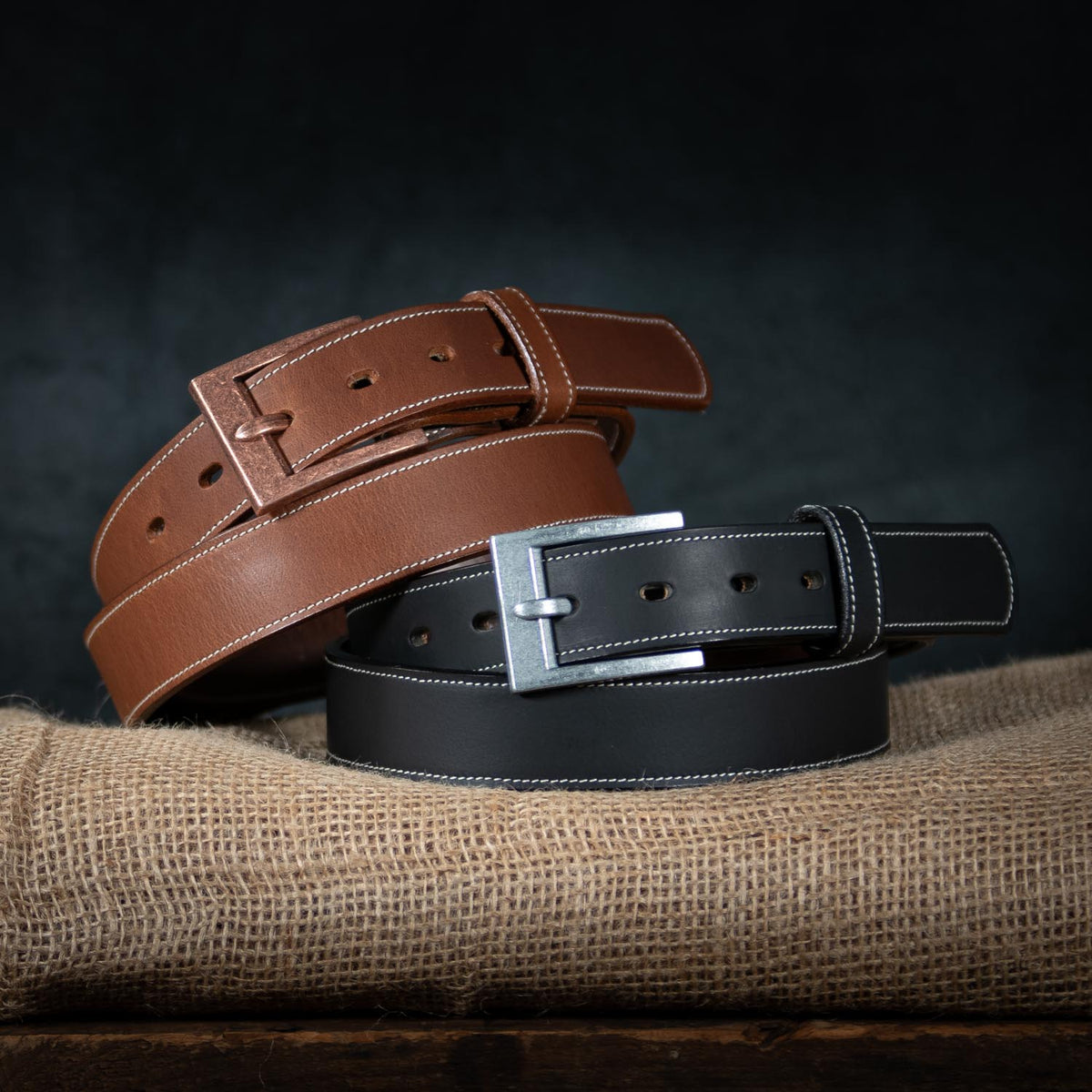 Two Hanks Belts "The Heritage Bison" leather belts—one brown, one black—feature white stitching and silver buckles. Displayed coiled on burlap, the brown belt sits atop the black, highlighting rugged style and American craftsmanship.
