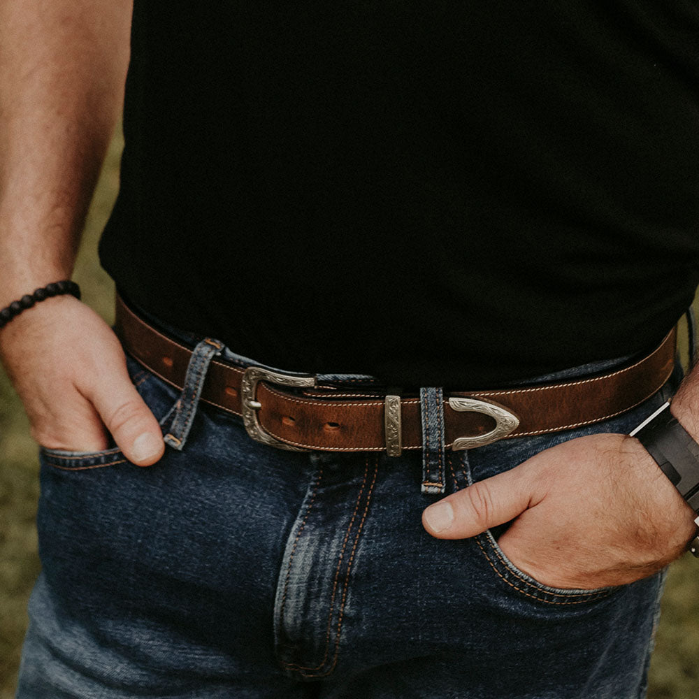 A person in a black shirt, blue jeans, and Hanks Belts' The Dakota Bison western belt with a floral buckle stands with hands in pockets. A black beaded bracelet and wristwatch complete the look, with grass visible in the background.