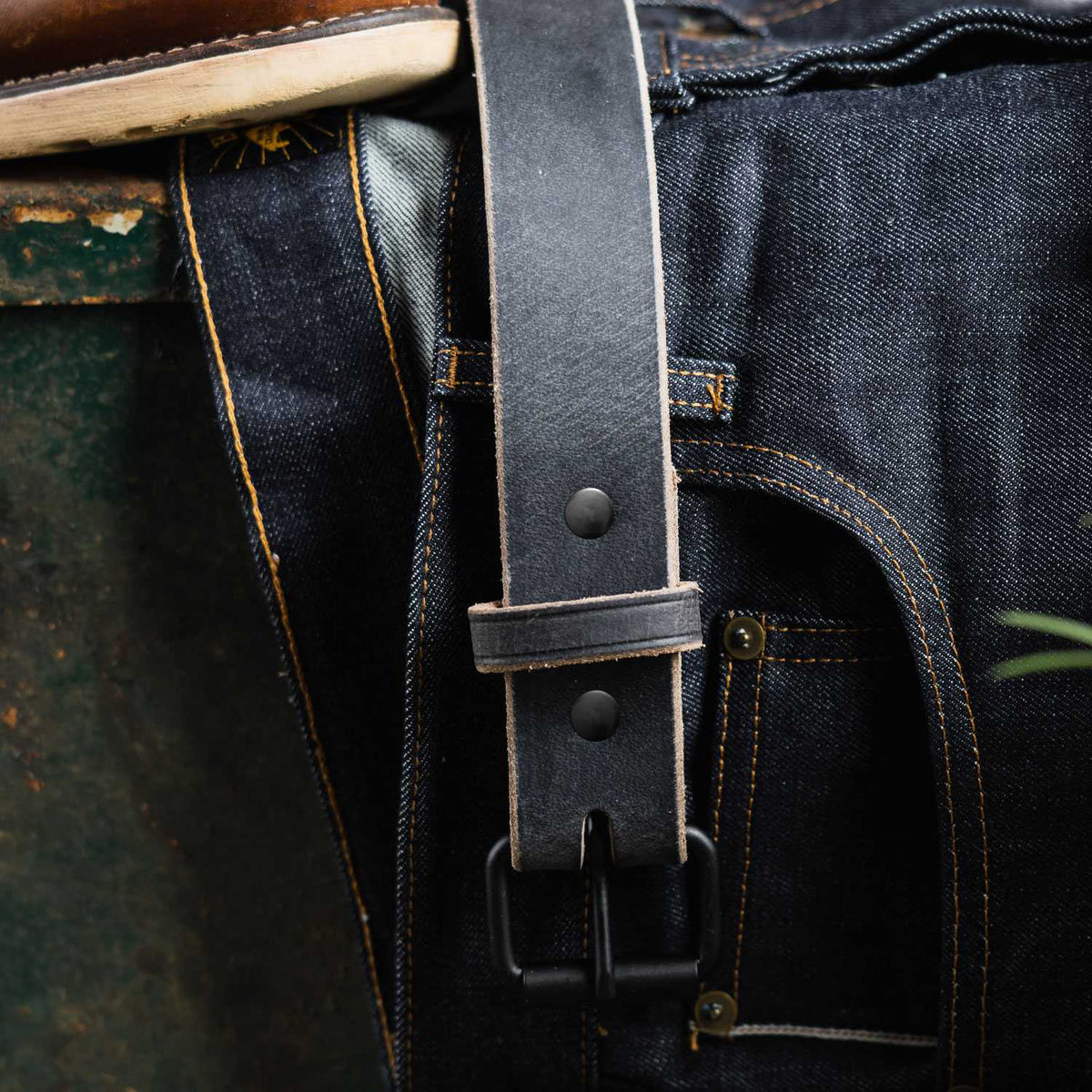 A close-up of Hanks Belts’ The Glacier Grey Retro Style, a water buffalo leather belt with a matte black buckle, draped over dark blue denim jeans with orange stitching beside a green textured surface.