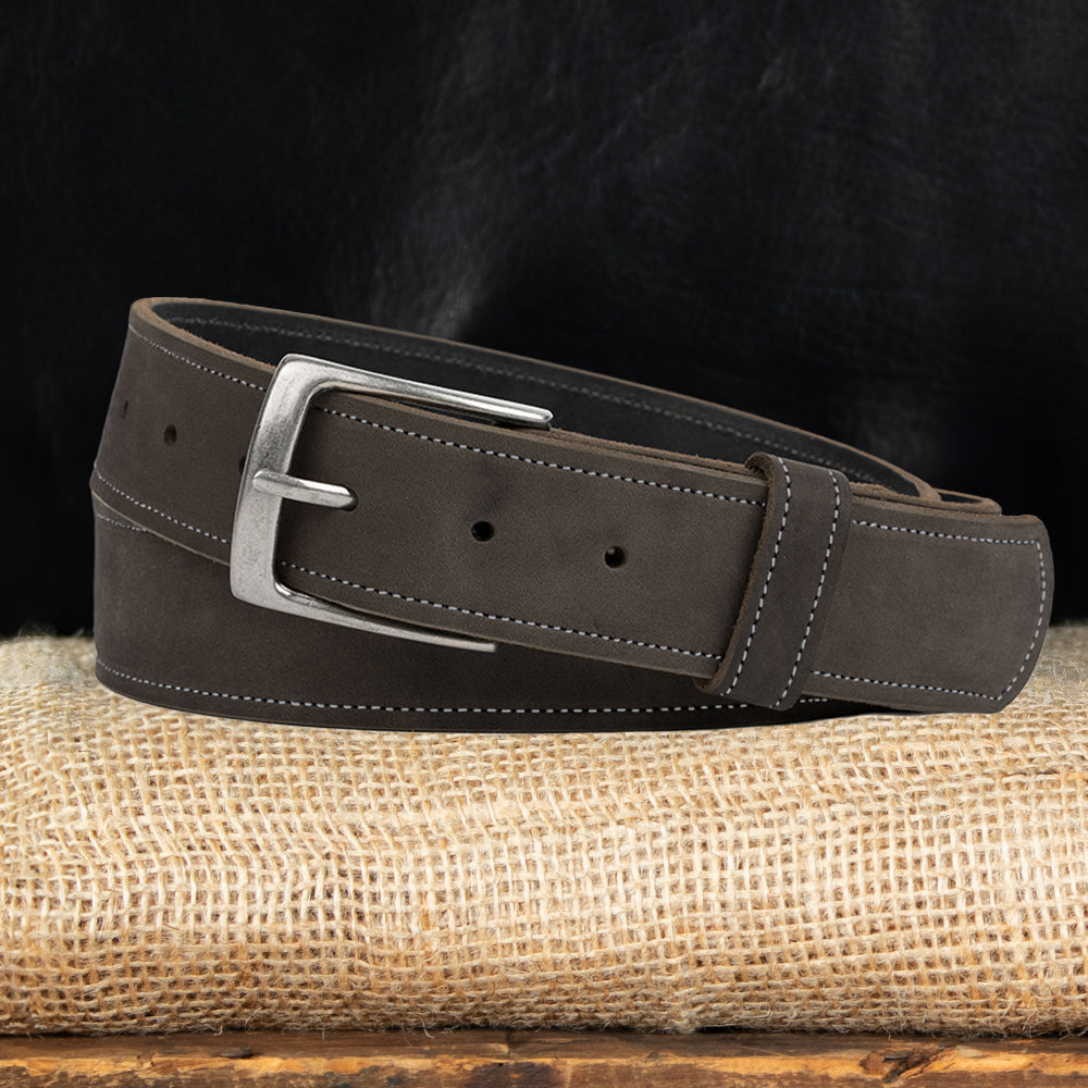 The Suede - 1.5" by Hanks Belts is a dark brown nubuck leather belt with white stitching and a silver buckle, shown coiled on burlap against a dark background. Proudly made in the USA.