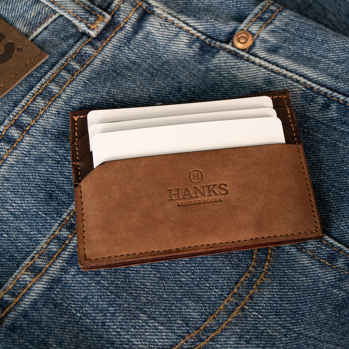 The Hanks Belts Whiskeytown Cardholder, in brown leather and holding white cards, rests on blue denim jeans, partially covering the back pocket and leather label.