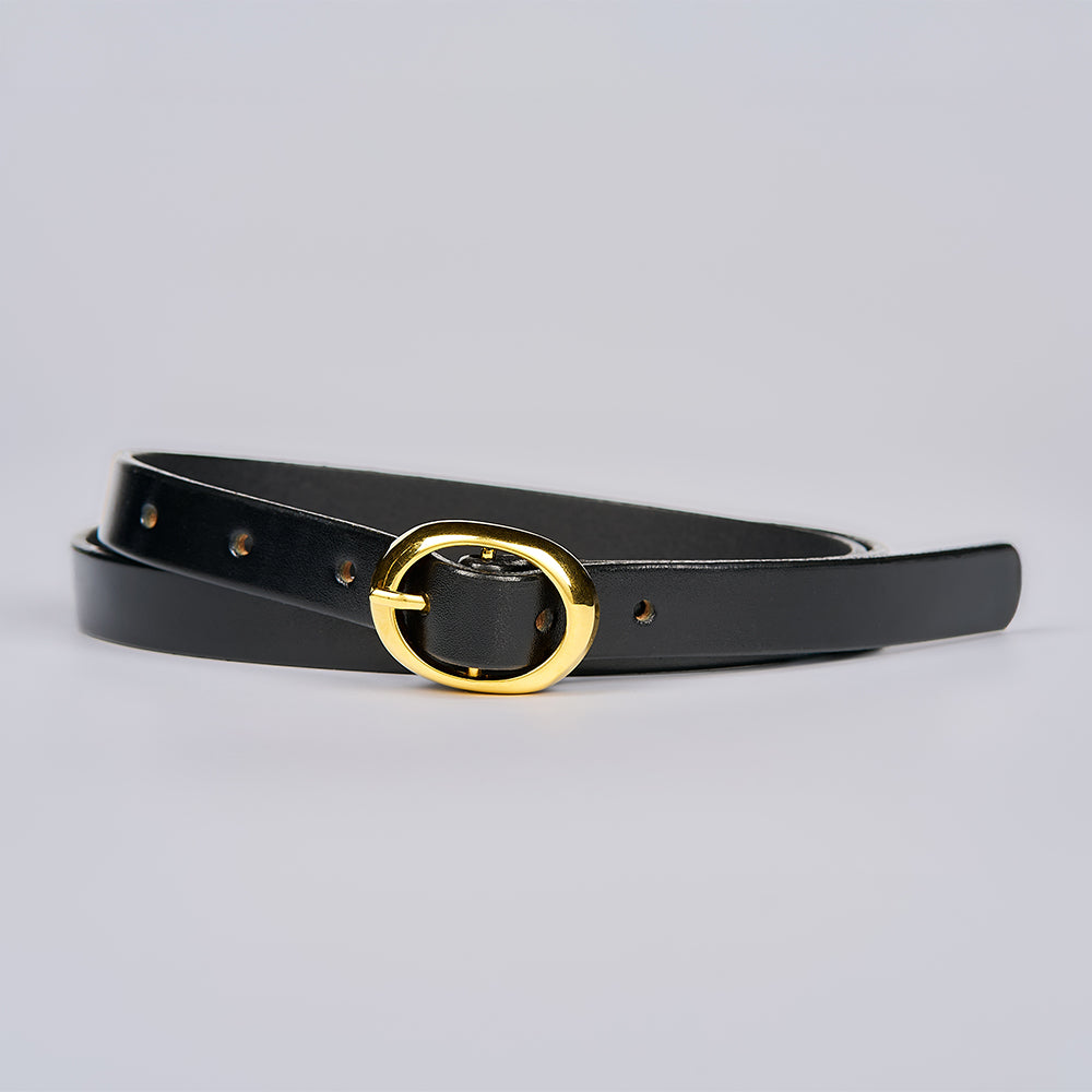 The Ivy by Hanks Belts, a black leather belt with a shiny gold oval buckle, is coiled on a plain light gray background.