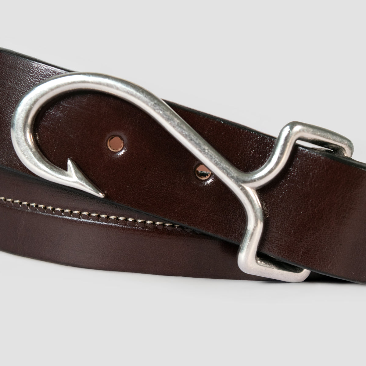 The Castaway - Limited Edition by Hanks Belts is a dark brown Havana English Bridle leather belt with a silver fish hook-shaped buckle, elegantly coiled on a light gray background.