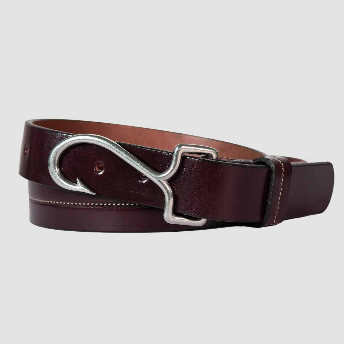 The Castaway - Limited Edition from Hanks Belts in Havana lies coiled on a light gray background, featuring English Bridle leather and a silver fish hook-shaped buckle.