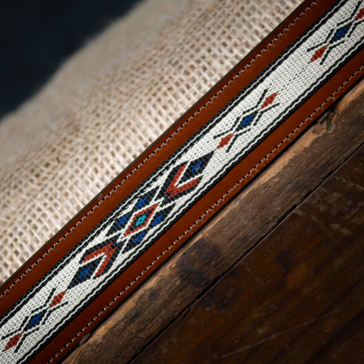 A close-up of The Seneca Tapered by Hanks Belts, made from premium English Bridle vegetable-tanned leather and featuring a red, black, blue, and white geometric embroidered pattern on a rustic wooden surface with burlap background.
