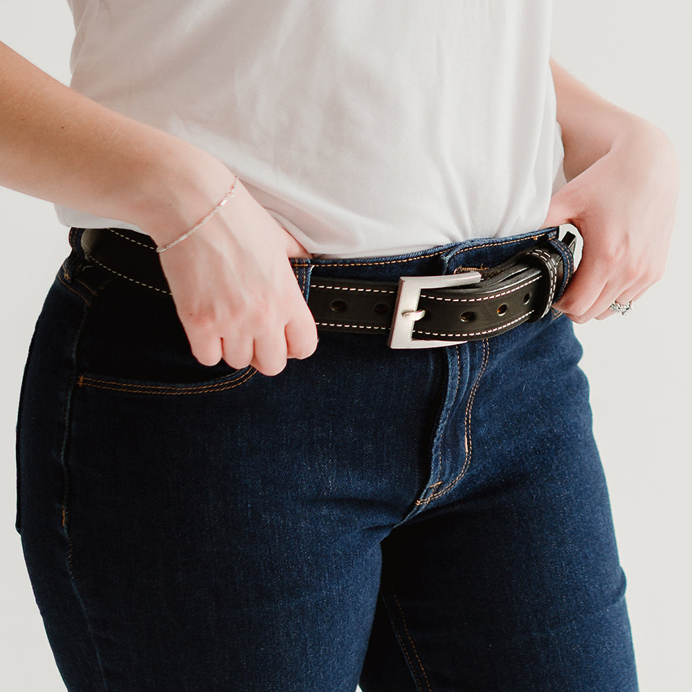A person in a white t-shirt and dark blue jeans adjusts The Bonnie full-grain vegetable-tanned leather belt with a silver buckle by Hanks Belts. Only their torso and upper legs are visible.