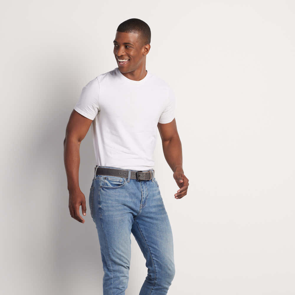 A man smiles to the side, wearing a white t-shirt, blue jeans, and The Glacier Grey Retro Style belt from Hanks Belts (USA Made), standing against a plain light background.