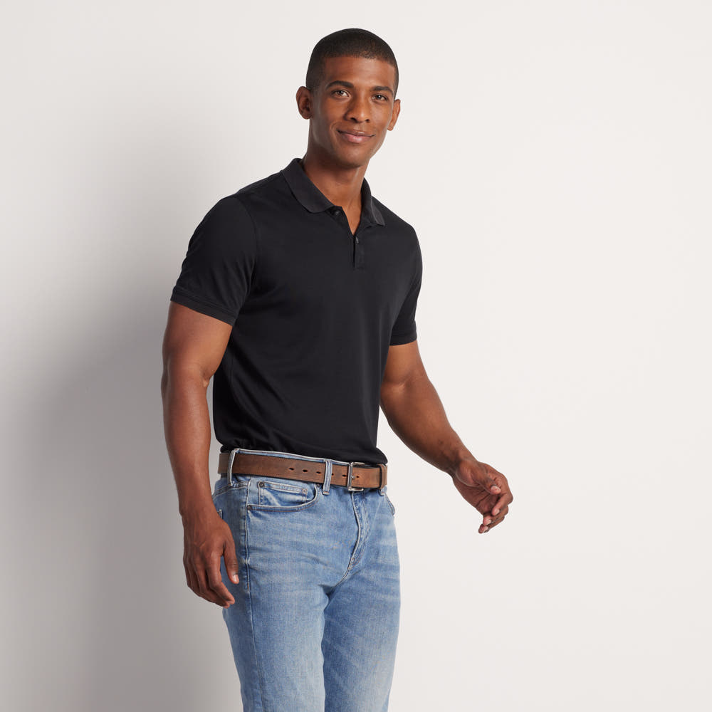 A man wearing a black short-sleeve polo, blue jeans, and the Hanks Belts Montana Bison Double Layer belt with a stainless steel buckle stands against a plain light background, smiling and looking slightly to the side.