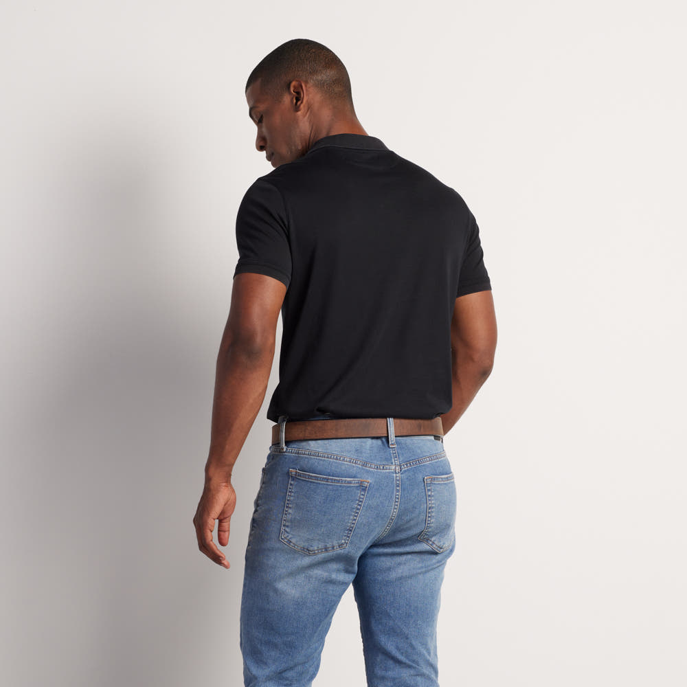 A man with short hair stands facing away, wearing a black polo shirt, light blue jeans, and the Hanks Belts "The Montana Bison Double Layer" rugged belt with a stainless steel buckle against a plain light background.