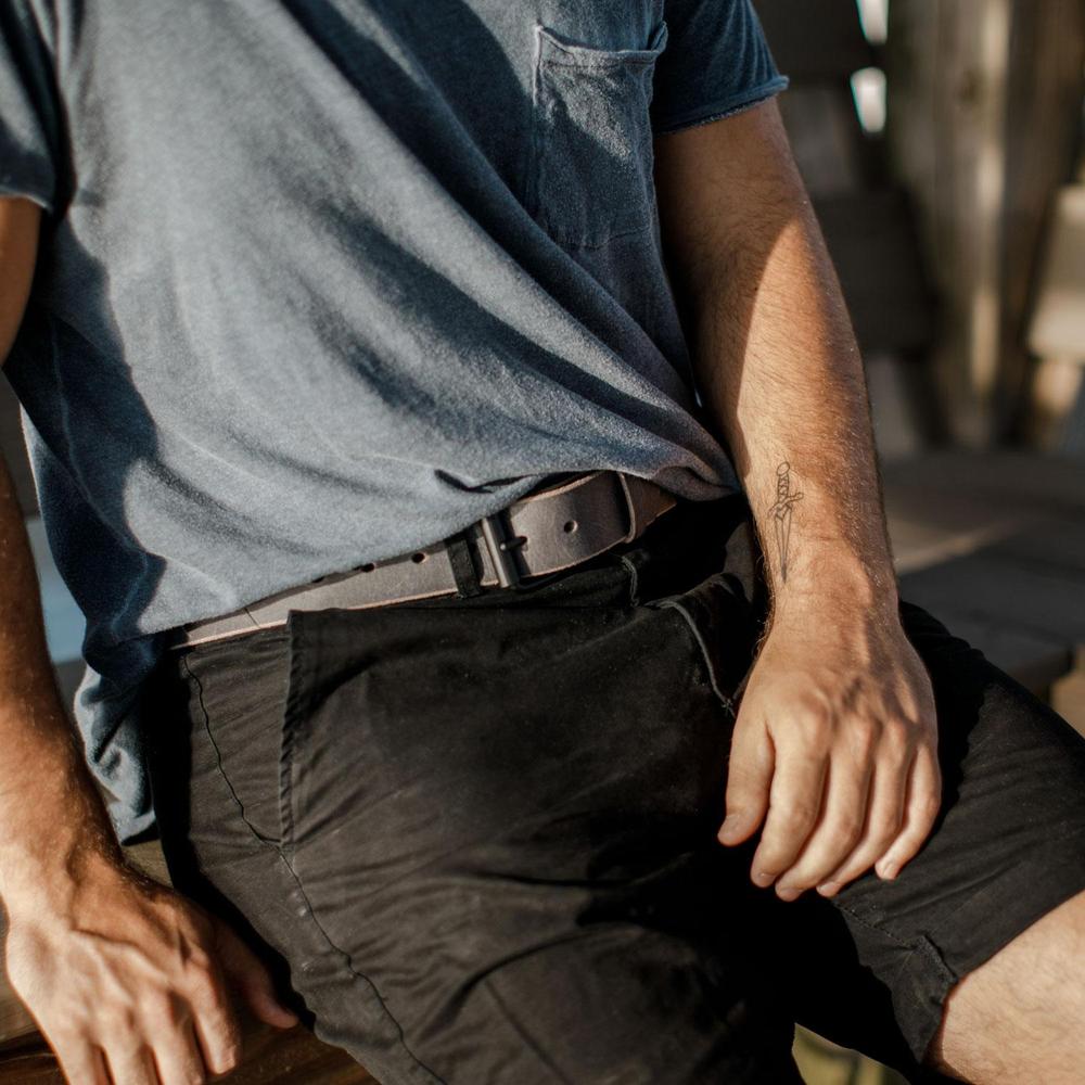 A person in a blue t-shirt and black shorts sits on wood, right arm on thigh. Their left forearm shows a sword tattoo, paired with The Glacier Grey Retro Style belt by Hanks Belts, crafted from USA made water buffalo leather.