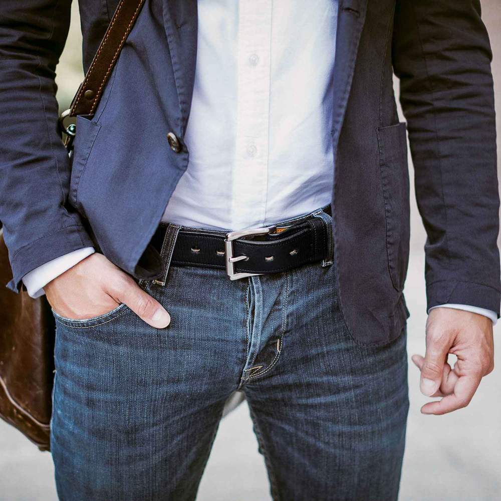 A person in a navy blazer, white shirt, and jeans wears The Tuscan Embossed Lined premium leather belt by Hanks Belts with a stainless steel buckle, one hand in their pocket and holding a brown shoulder bag.