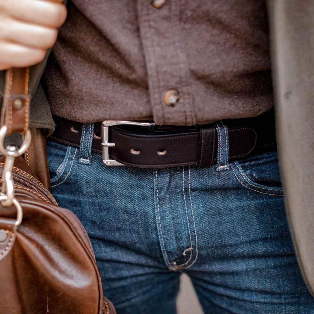 A person in a brown button-up shirt and blue jeans wears the Hanks Belts "The Tuscan Embossed Lined" premium leather belt with a stainless steel buckle, paired with a brown leather bag.