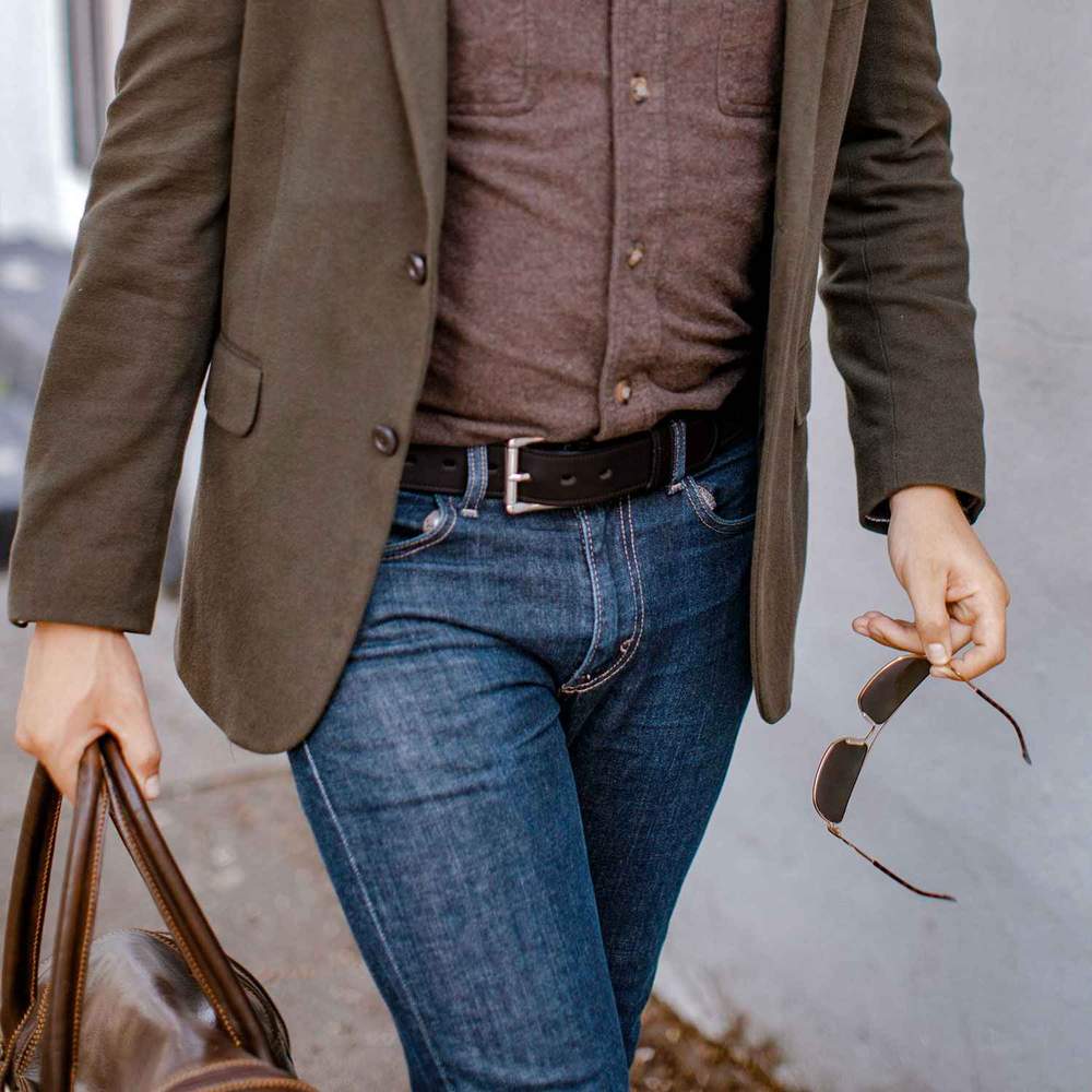 A person walks outdoors in a brown blazer and shirt, blue jeans, holding sunglasses and a brown leather bag, accessorized with Hanks Belts' The Tuscan Embossed Lined premium leather belt with stainless steel buckle.