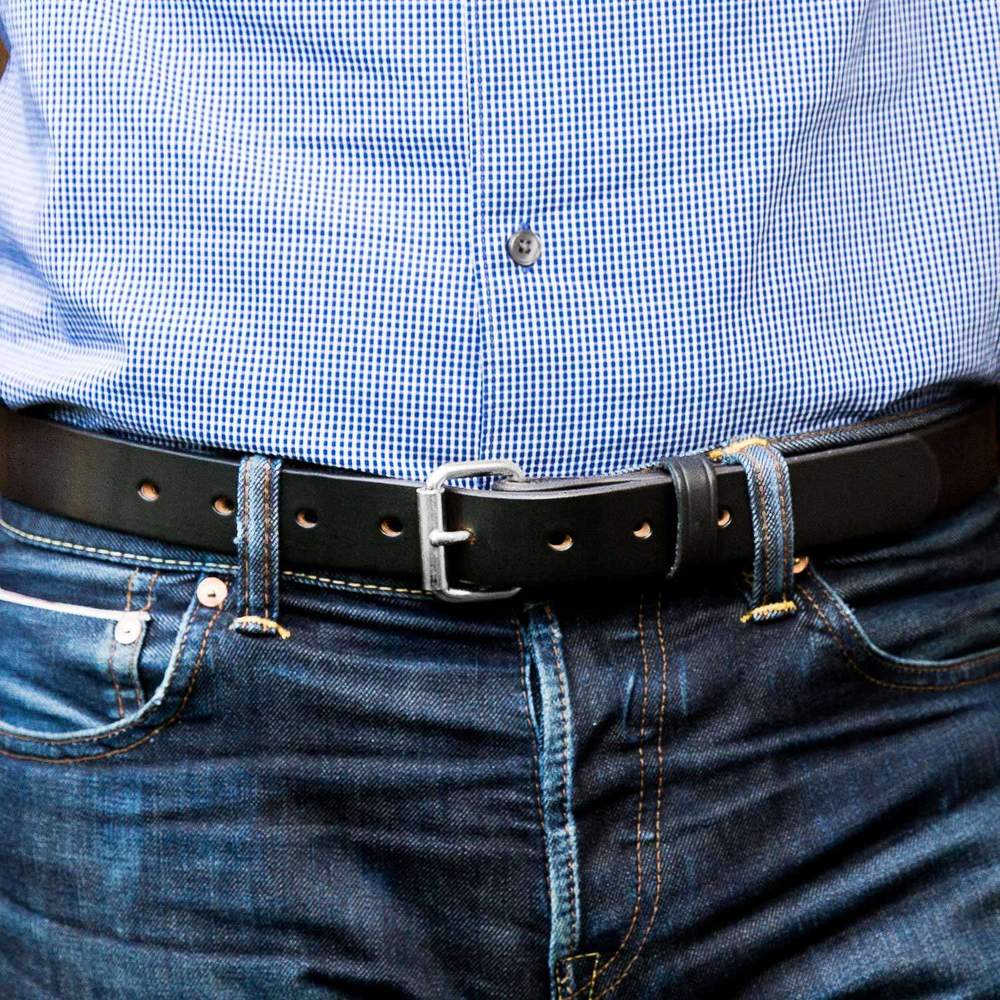 Close-up of a person wearing a blue checkered shirt tucked into blue jeans, secured with The Deputy by Hanks Belts—a durable concealed carry belt with a black leather strap and silver buckle.