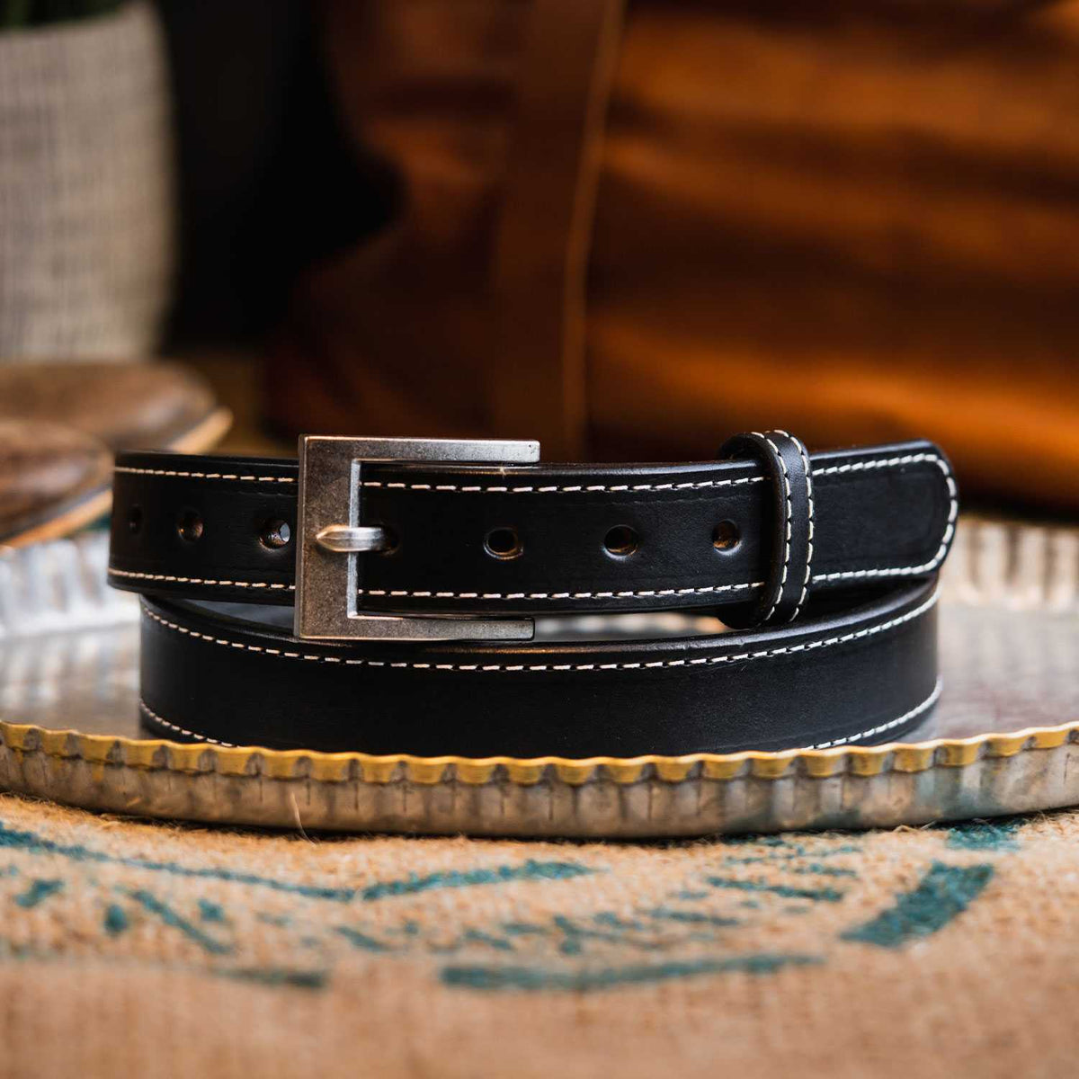 The Bonnie by Hanks Belts, a black full-grain vegetable tanned leather belt with white stitching and a silver buckle, is coiled on a metal tray against a blurred brown and beige background.