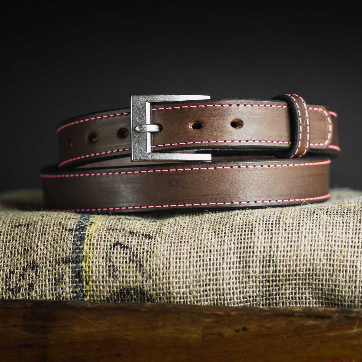 The Bonnie by Hanks Belts, made from full-grain vegetable-tanned leather with pink stitching and a silver buckle, is coiled on burlap against a dark background.