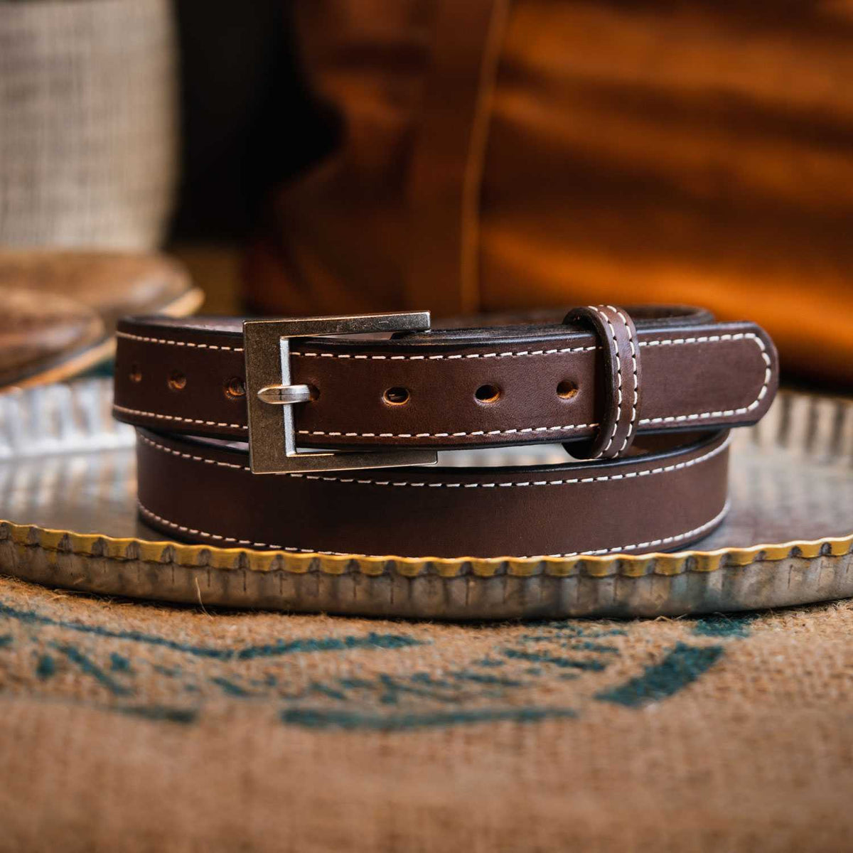 A coiled The Bonnie belt from Hanks Belts, made of full-grain vegetable tanned leather with white stitching and a silver buckle, rests on a metal tray atop burlap.