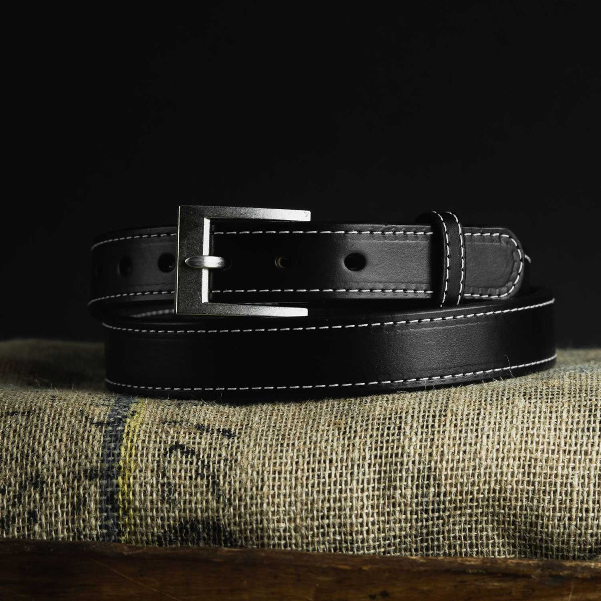 The Bonnie by Hanks Belts, made from full-grain vegetable-tanned leather with white stitching and a silver buckle, is coiled on textured burlap against a black background.