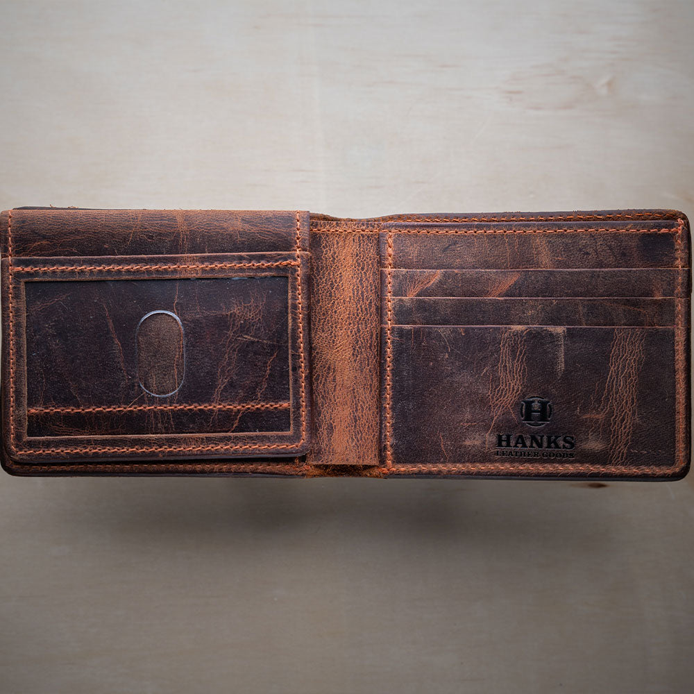 The Powell Bison Bifold with ID Window Flap by Hanks Belts is shown open on a light surface, revealing card slots, an ID window, and the "Hanks" logo embossed inside its worn brown leather.