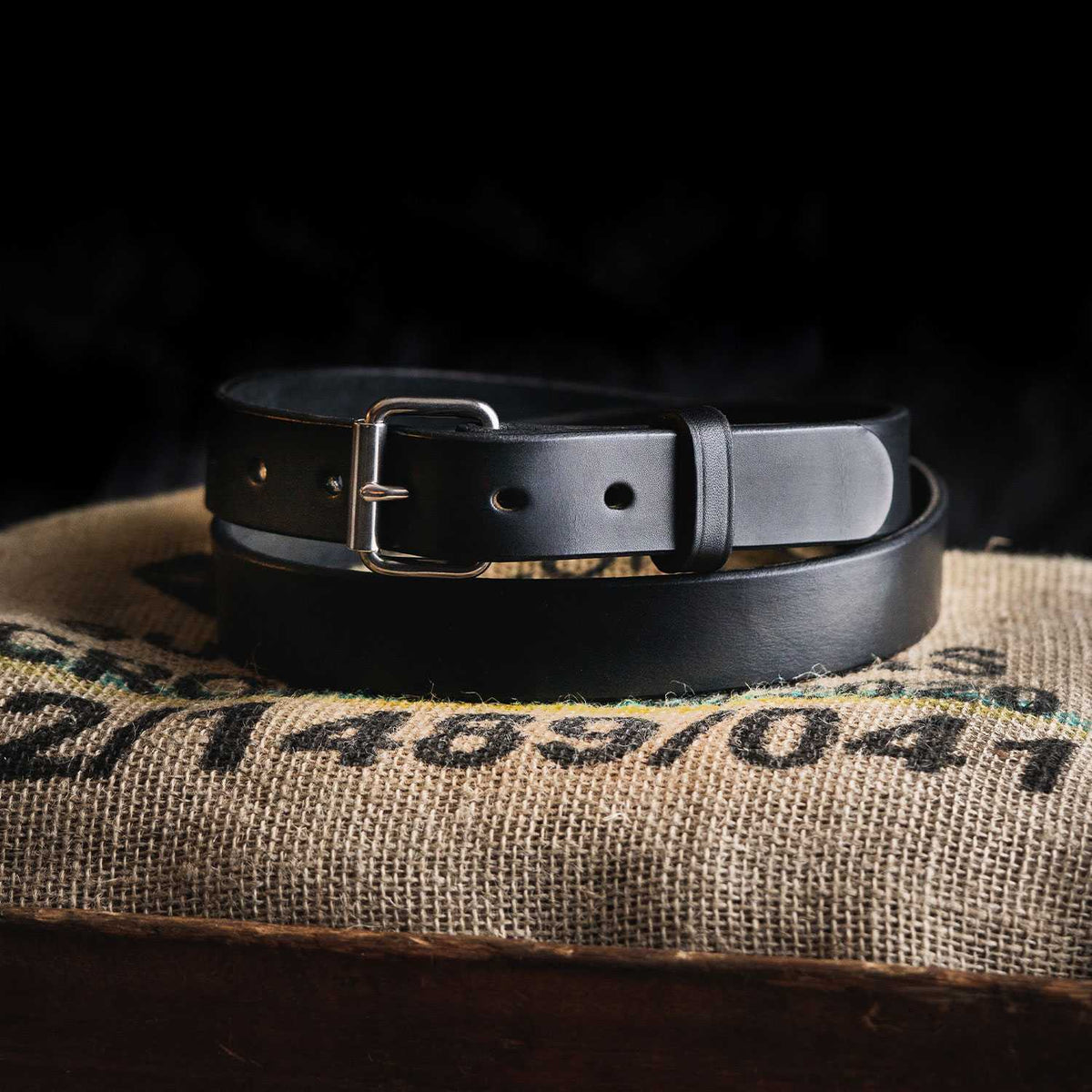 The Deputy leather belt by Hanks Belts, featuring a silver buckle, is coiled on a numbered burlap sack against a dark background.