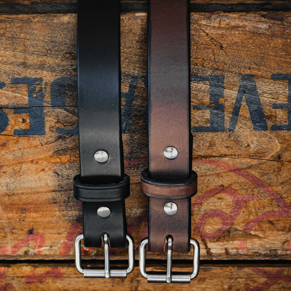 The Deputy by Hanks Belts: two leather belts, black and brown with silver buckles and rivets, displayed side by side on a rustic wooden surface—perfect for durable daily wear.