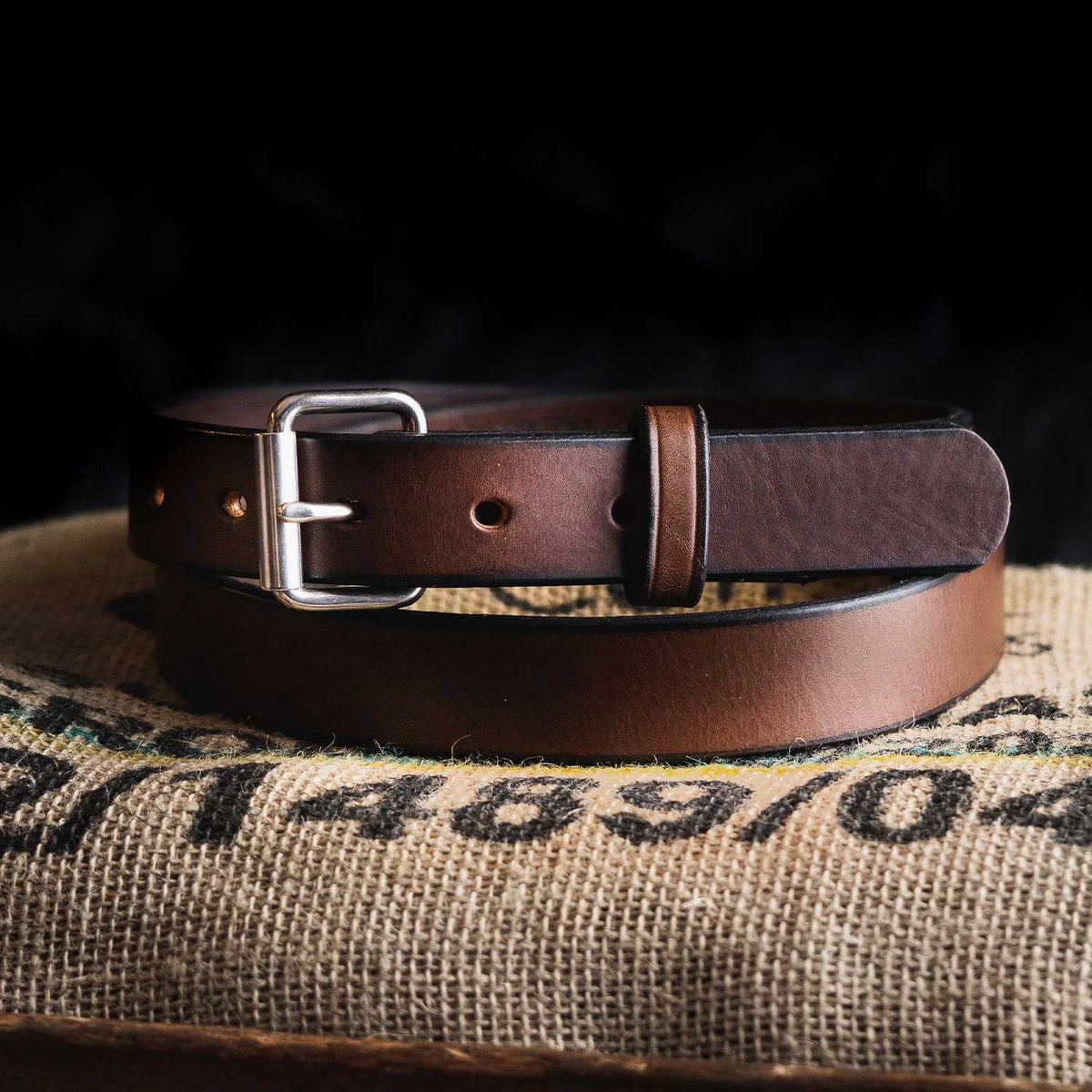 The Deputy leather belt by Hanks Belts, featuring a silver buckle, is coiled on a burlap sack with subtle black and green markings, set against a dark background.
