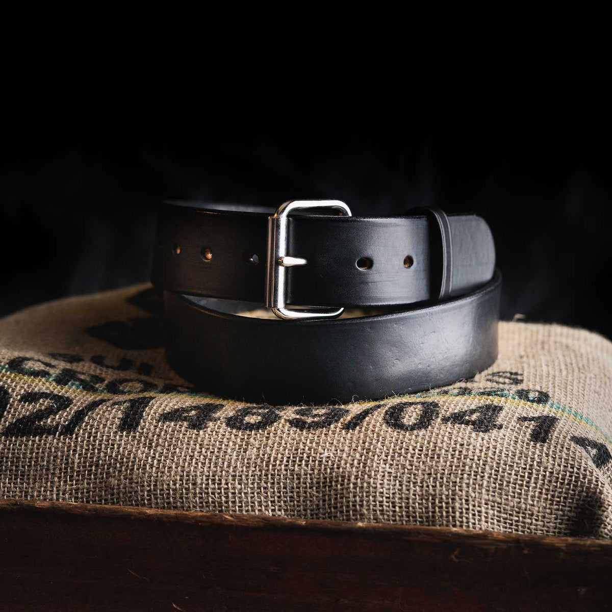 The Enforcer full-grain leather belt from Hanks Belts, featuring a silver buckle, is coiled neatly on a burlap sack with black and green text against a dark background.