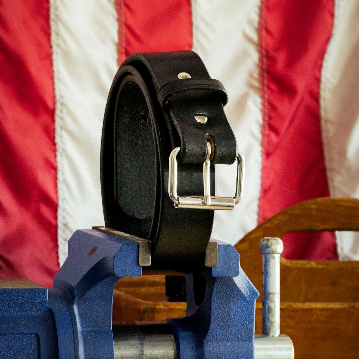 Hanks Belts' The Enforcer, a full-grain leather belt with a silver buckle, is clamped in a blue vise, with part of a wooden chair and an American flag in the background.