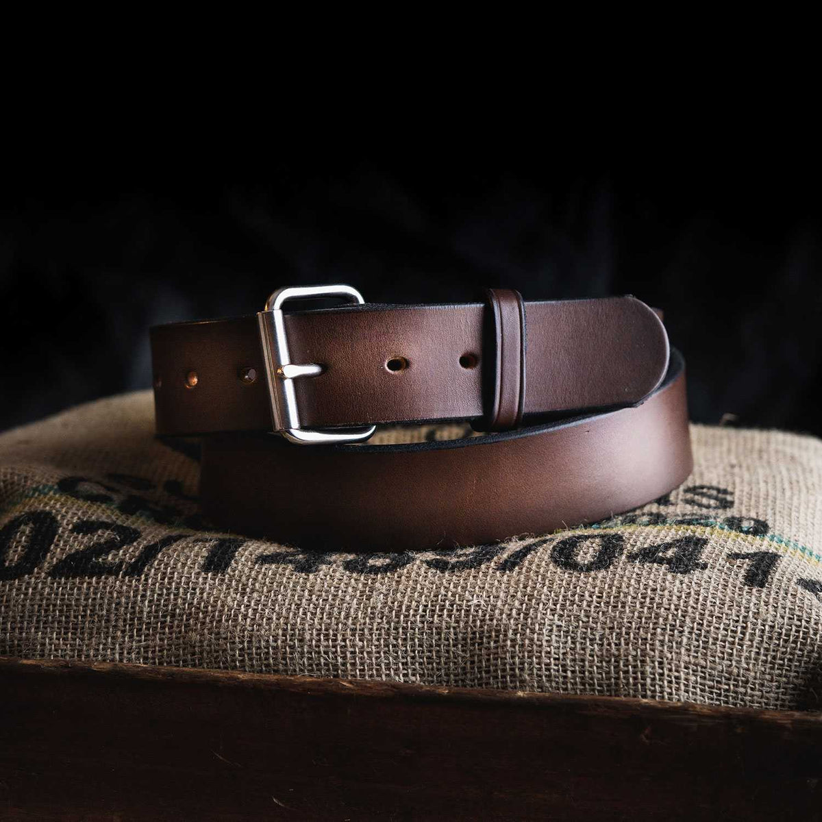 The Enforcer full-grain leather belt by Hanks Belts features a silver buckle and is rolled up on a burlap-covered surface with black text, set against a dark background.