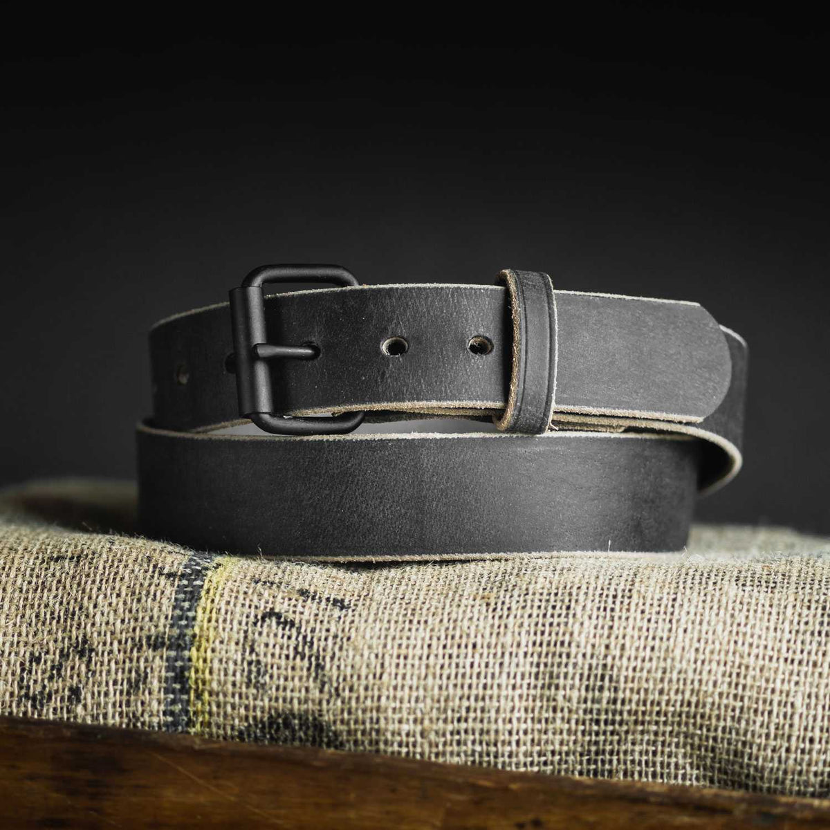 The Glacier Grey Retro Style belt by Hanks Belts, made in the USA from durable Water Buffalo leather with a matte black buckle, is coiled neatly on burlap fabric against a dark background.