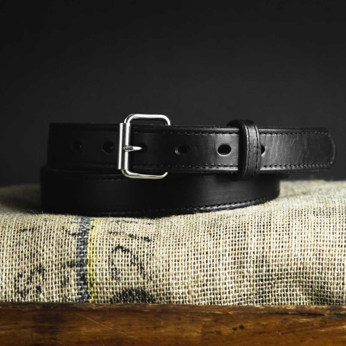 The Hanks Belts "The Steel Core," a black leather gun belt with a silver buckle, is neatly coiled and displayed on textured beige cloth against a dark background.