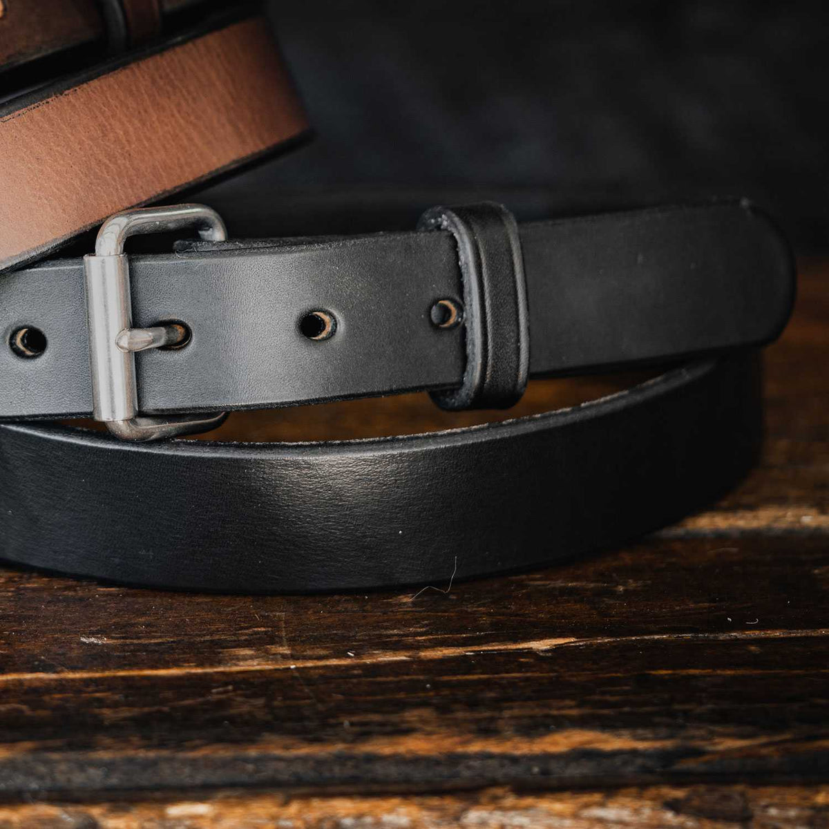 Close-up of Hanks Belts' "The Deputy" leather concealed carry belt in black with a metal buckle, prominently displayed in the foreground; a brown version is partially visible behind it on a wooden surface.