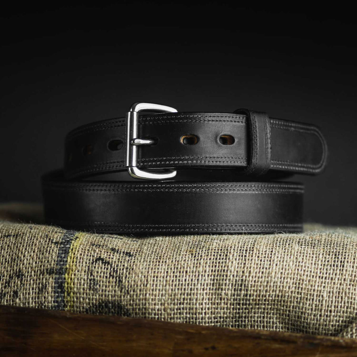 The Hanks Belts Old World Harness Leather Belt - 1.5", crafted from USA-tanned Hermann Oak black full-grain leather with a silver buckle, is coiled on burlap against a dark background.