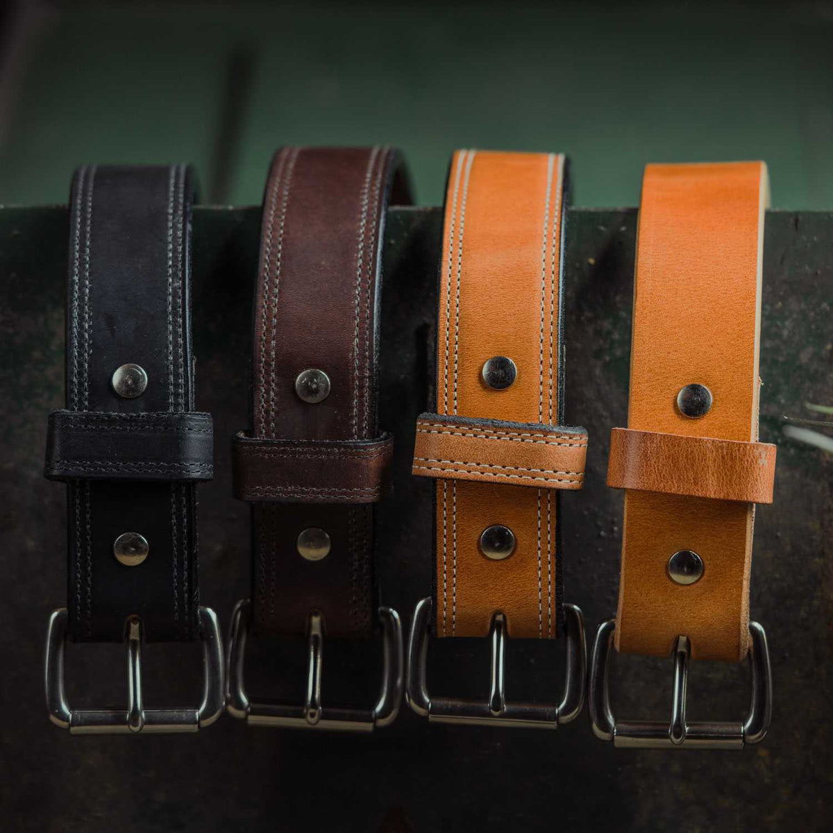Four Hanks Belts Old World Harness Leather Belts (1.5") in black, dark brown, tan, and light brown hang side by side on a dark surface, each crafted from full-grain leather with metal buckles and visible stitching.