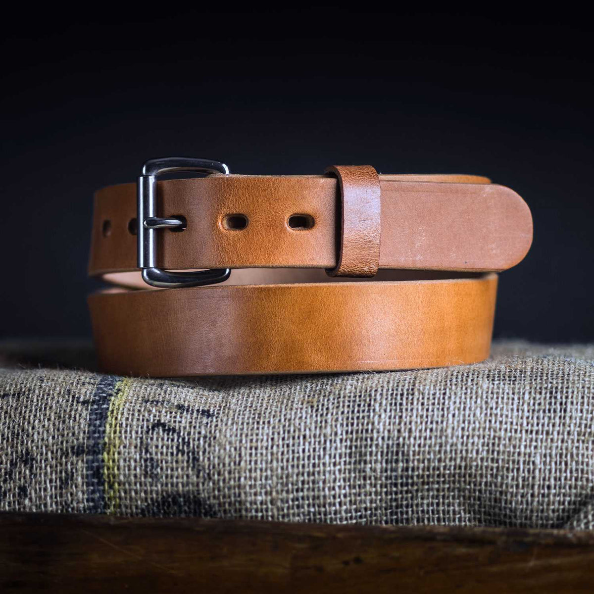 The Old World Harness by Hanks Belts, a tan full-grain leather belt with a silver buckle, is coiled on a burlap surface against a dark backdrop.