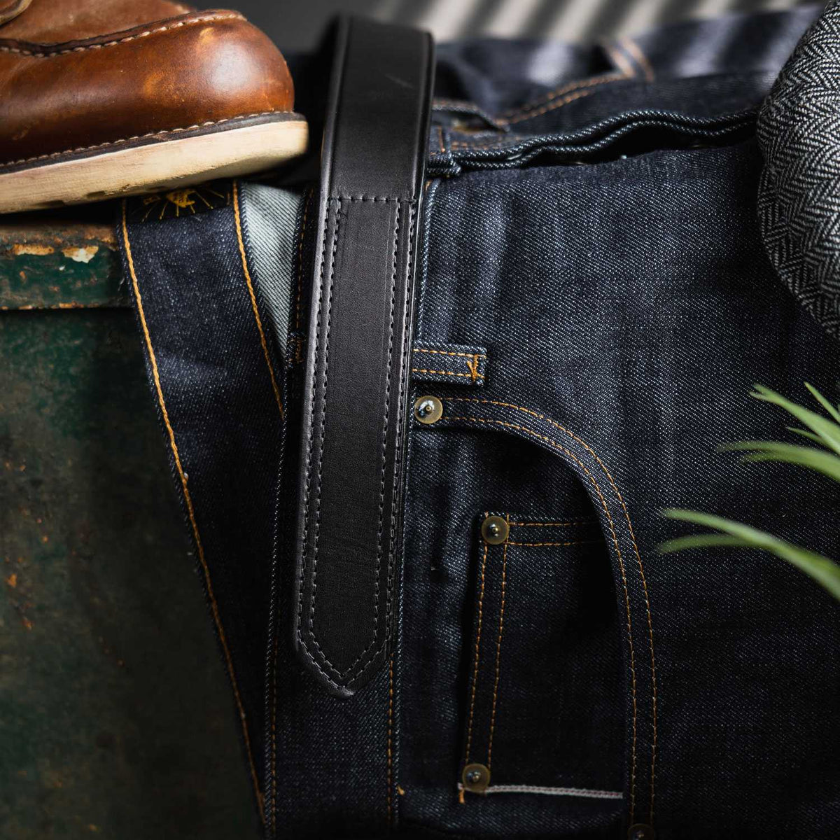 A close-up shows dark blue jeans with The Scratchless Mechanics black belt by Hanks Belts threaded through the loops, next to a brown boot on textured gray fabric. A green plant is partially visible in the foreground.