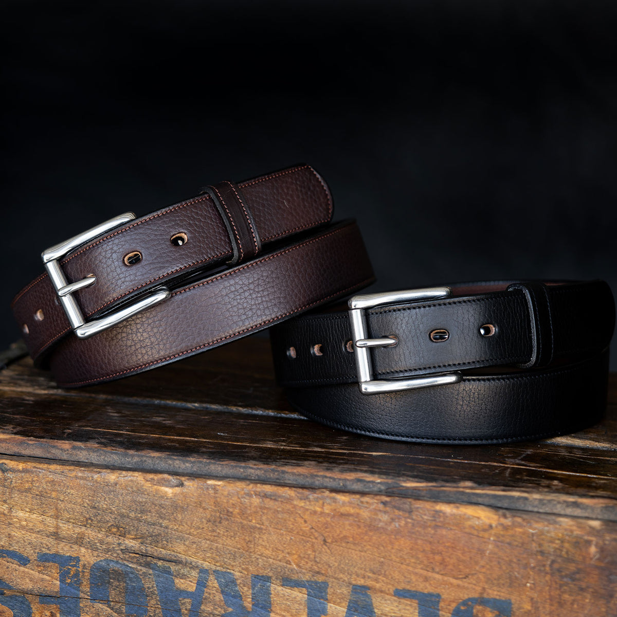 Two Hanks Belts "The Tuscan Embossed Lined," one brown and one black, each featuring a stainless steel buckle, are displayed side by side on a rustic wooden surface against a dark backdrop.